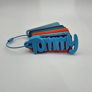 May include: A set of stacked, multi-colored rectangular luggage tags held together by a blue wire cable. The top tag is light blue and features the name Tommy in raised, cursive lettering. The tags underneath are teal, purple, orange, and red.