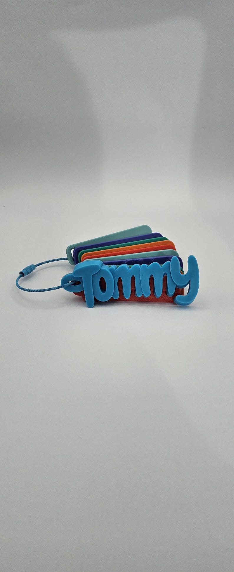 May include: A set of stacked, multi-colored rectangular luggage tags held together by a blue wire cable. The top tag is light blue and features the name Tommy in raised, cursive lettering. The tags underneath are teal, purple, orange, and red.