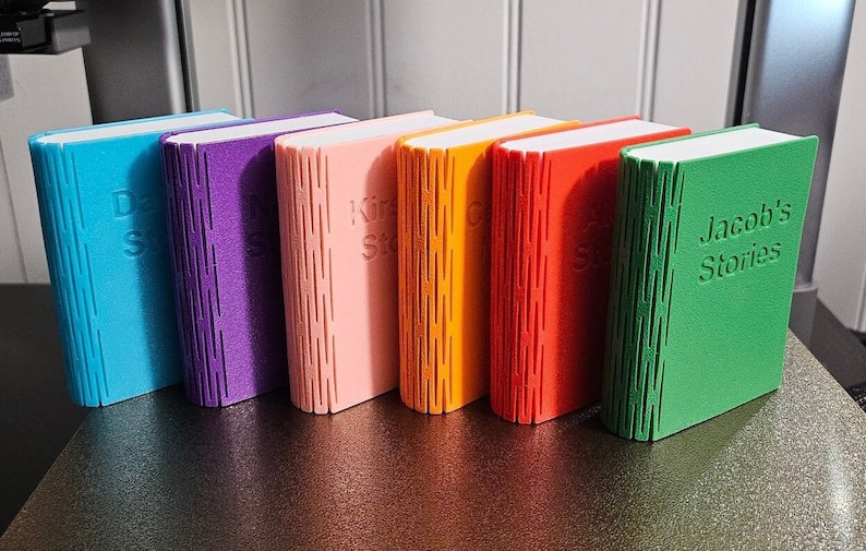 Selection of 3d printed storage boxes for holding Yoto cards. They are designed to look like a book, have a colour cover, white pages, and can be customised with any text.