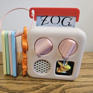 May include: A white square electronic toy with two pink knobs, a speaker grille, and a small screen displaying a pixelated yellow creature. A red tab at the top holds a card labeled ZOG. A stack of pastel-colored plastic cards is attached by a wire loop.