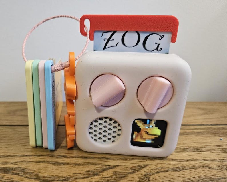 May include: A white square electronic toy with two pink knobs, a speaker grille, and a small screen displaying a pixelated yellow creature. A red tab at the top holds a card labeled ZOG. A stack of pastel-colored plastic cards is attached by a wire loop.