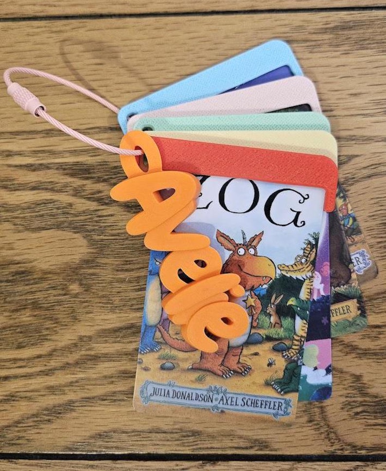 May include: A set of stacked, rectangular plastic cards featuring illustrations from the book Zog by Julia Donaldson and Axel Scheffler. The cards are held together by a pink wire loop and a bright orange 3D-printed name tag reading Janelle.