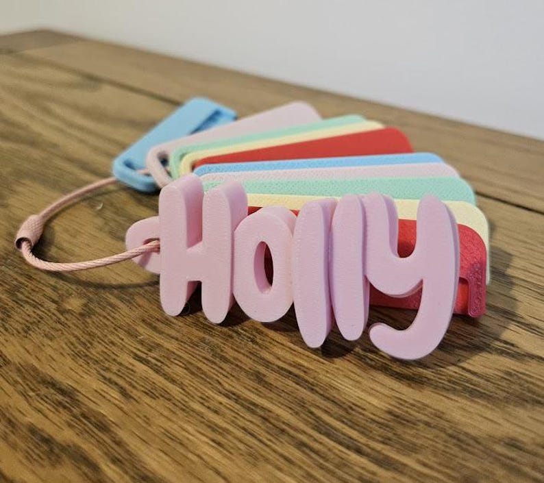 May include: A fan-style collection of 3D-printed plastic name tags, each featuring a different name in a rounded, bold font. The tags are attached to a thin, flexible pink cord. The colors include light pink, blue, mint green, yellow, and red.