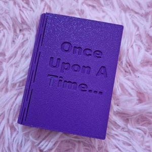 K&ouml;nnte beinhalten: Ein rechteckiges, violettes Objekt in Form eines kleinen Buches mit strukturierter Oberfl&auml;che. Auf dem Vorderdeckel befindet sich der gepr&auml;gte Text "Once Upon A Time..." in einer serifenlosen Schrift sowie vertikale Linien am Buchr&uuml;cken.