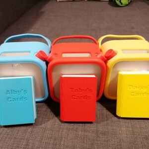 K&ouml;nnte beinhalten: Drei quadratische Beh&auml;lter mit Griff in Hellblau, Rot und Gelb, jeweils mit einem passenden kleinen buchf&ouml;rmigen Objekt davor. Das blaue Buch ist mit Alby's Cards gepr&auml;gt, das rote mit Toby's Cards und das gelbe mit Daisy's Cards.