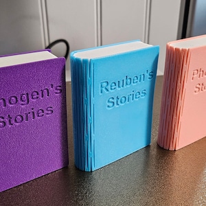 3d printed storage boxes for holding Yoto cards. Designed to look like a book, these ones are Pink, Purple and Blue with white pages and has each has a customised cover bearing the name of the owner. This can be fully customised.