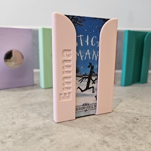 May include: A light pink plastic holder with the name Emma embossed on the side, containing a book titled Stick Man by Julia Donaldson and Axel Scheffler. Additional holders in lavender, light green, and teal are visible in the background.