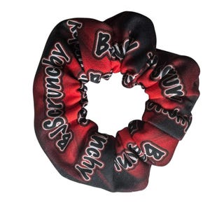 May include: A red and black scrunchy with the words "Ba Scrunchy" in white script. The scrunchy is made of a soft fabric and is designed to hold hair in place. The scrunchy is a hair accessory.