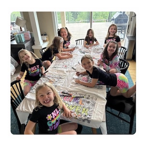 May include: A group of girls are gathered around a table, coloring a large canvas with the words "GIRL POWER" and other designs. They are wearing black t-shirts with colorful graphics and tie-dye shorts. The scene is set indoors with natural light.