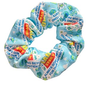 May include: A light blue scrunchie with a repeating pattern of cartoon characters, stars, and the words "Toy Story" and "Always Going Beyond for Our Patients" in red, yellow, and white. The scrunchie is made of a soft, gathered fabric.