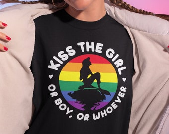 May include: Black t-shirt with a circular graphic featuring a mermaid silhouette on a rainbow background. The text reads "KISS THE GIRL, OR BOY, OR WHOEVER" in a white, bold font. The shirt is worn by a person with braided hair and red lipstick.
