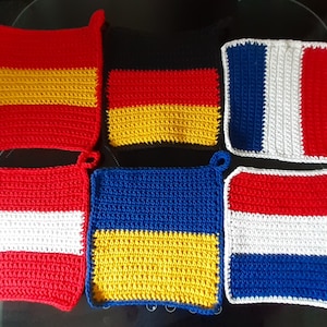 Puede incluir: Seis agarraderas de olla de ganchillo, cada una con los colores de la bandera de un país diferente. Las banderas son las de España, Alemania, Francia, Austria, Ucrania y los Países Bajos.