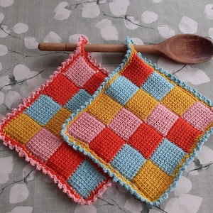 Può includere: Due presine quadrate all'uncinetto con un motivo a scacchi multicolore nelle tonalità del rosso, azzurro, giallo e rosa. Ogni presina presenta un bordo decorativo smerlato ed è posizionata vicino al manico di un cucchiaio di legno.