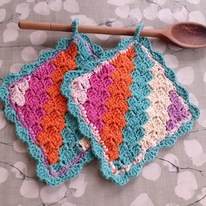 Könnte beinhalten: Zwei gehäkelte Topflappen mit einem gewellten Rand. Die Topflappen sind aus einem mehrfarbigen Garn in den Farben Pink, Orange, Türkis und Creme gefertigt.