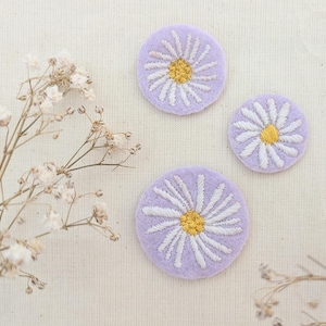 May include: Three embroidered daisy patches in different sizes. Each patch has a lavender background, white petals, and a yellow centre. They are arranged on cream-coloured fabric, with dried flowers in the background.