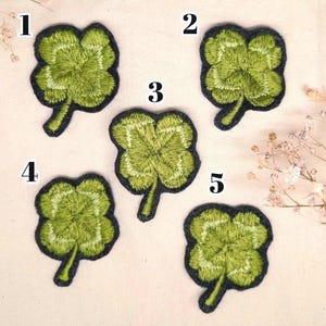 May include: Five embroidered four-leaf clover patches. Each clover is a vibrant green with detailed stitching and a black border. The patches are arranged on a light-coloured fabric, with delicate white flowers in the background.
