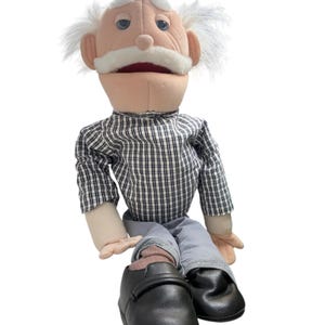 May include: A plush puppet of an elderly person with white hair and a white mustache, wearing a black and white checkered long-sleeve shirt, light gray trousers, and black shoes. The puppet has a light skin tone, blue eyes, and a red mouth interior.