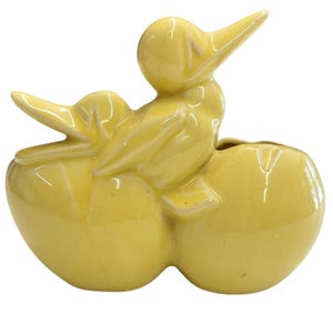 May include: A glossy yellow ceramic planter shaped like a duck sitting atop a large, split egg. The duck has a smooth, rounded head, a pointed beak, and wings tucked against its body. The base consists of two rounded, bulbous sections.