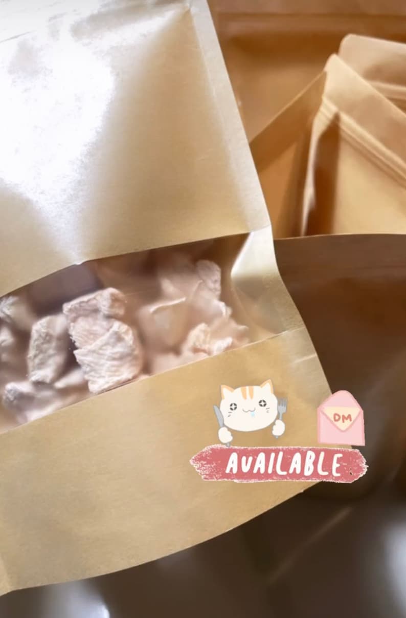 Puede incluir: Un primer plano de una bolsa de papel marr&oacute;n con una ventana transparente que muestra malvaviscos blancos y esponjosos en su interior. La bolsa tiene una pegatina con un gato de dibujos animados sosteniendo un cuchillo y un tenedor, y el texto "AVAILABLE" en rosa.