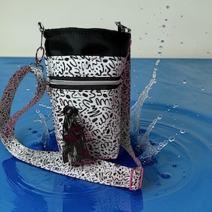 May include: A black and white patterned dog waste bag holder with a matching strap. The bag holder has a black top and a zippered pocket. A roll of waste bags is attached to the front. The strap has pink accents.