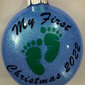 Puede incluir: Adorno navideño azul con purpurina, con huellas verdes y el texto "My First Christmas 2022".