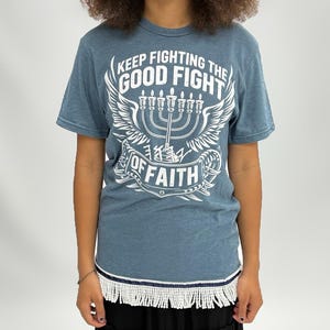 May include: A blue t-shirt with the text "KEEP FIGHTING THE GOOD FIGHT OF FAITH" and a menorah graphic. The shirt has a fringe trim at the bottom. The person in the image has curly brown hair with red highlights.