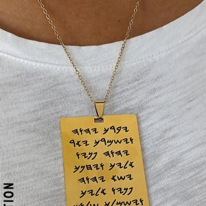 Könnte beinhalten: Goldfarbene rechteckige Anhängerhalskette mit feiner Kette. Der Anhänger hat eingravierten Text in einer stilisierten Schriftart. Die Halskette wird über einem weißen T-Shirt getragen. Der Markenname "X NATION" ist links aufgedruckt.