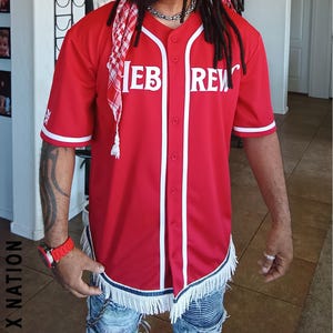 May include: Red baseball jersey with white trim and the text "HEB REW" in white block letters. The jersey has a button-up front and short sleeves. The bottom hem is decorated with white fringe. The person is wearing a red and white headscarf.