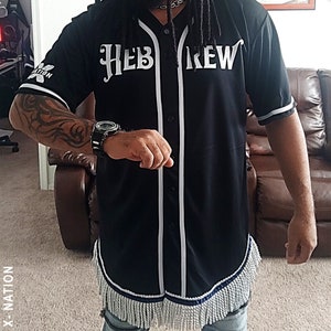 May include: A black baseball jersey with white trim and the word "HEBREW" in white lettering. The jersey has a fringe trim at the bottom and is worn by a person with long dreadlocks and a bandana.