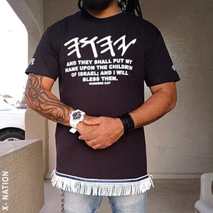 以下が含まれることがあります： 白いヘブライ語のテキストと英語の引用句が書かれた黒いTシャツ: "AND THEY SHALL PUT MY NAME UPON THE CHILDREN OF ISRAEL; AND I WILL BLESS THEM." シャツの裾には白と青のフリンジが付いています。画像には、白黒のヘッドラップも表示されています。