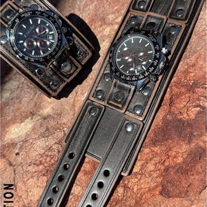 May include: Two black and silver wristwatches with black leather bands. The watch faces display the text "X NATION" and have multiple sub-dials. The leather bands are wide and feature metal rivets and decorative stitching. The watches are displayed on a textured, reddish-brown surface.