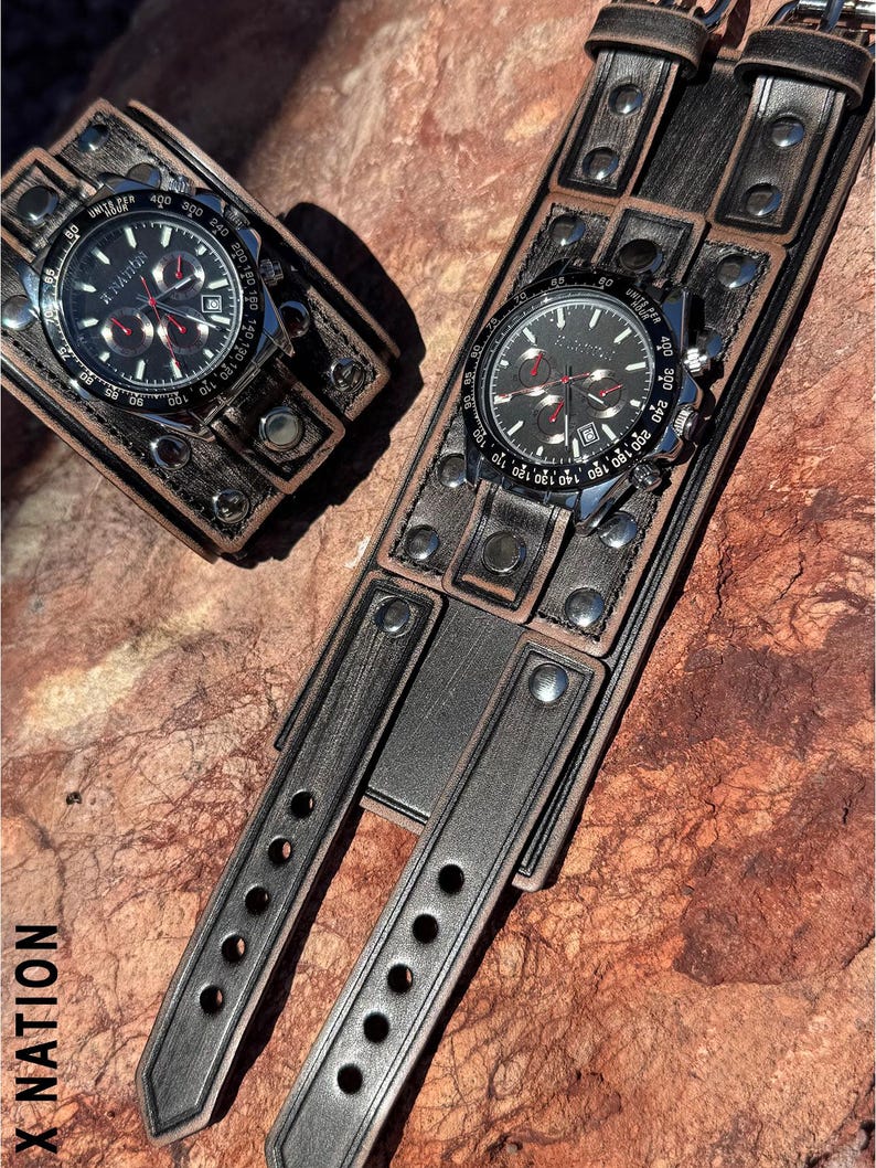 May include: Two black and silver wristwatches with black leather bands. The watch faces display the text "X NATION" and have multiple sub-dials. The leather bands are wide and feature metal rivets and decorative stitching. The watches are displayed on a textured, reddish-brown surface.