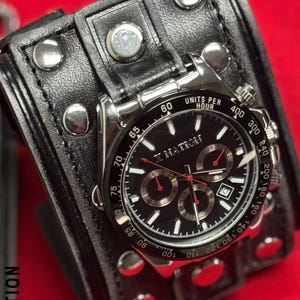 May include: A black wristwatch with a silver bezel and a black leather cuff. The watch face has white and red accents, with multiple sub-dials. The cuff is studded with silver rivets and has the text "X NATION".