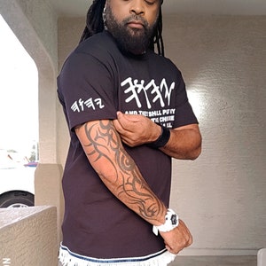 May include: Black t-shirt with white Hebrew text and fringe trim. The shirt has a crew neck and short sleeves. The text on the shirt reads "AND THE SHALL PUTY THE CHINE". The image also shows a black and white head wrap and a white watch.