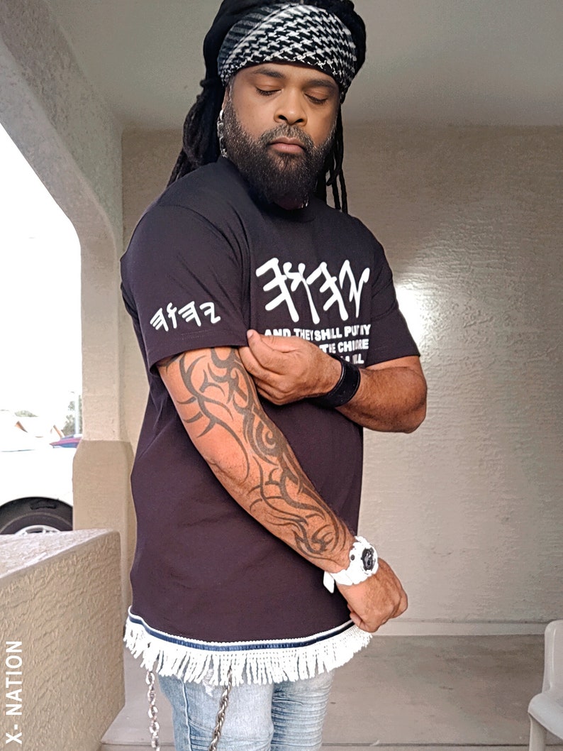 May include: Black t-shirt with white Hebrew text and fringe trim. The shirt has a crew neck and short sleeves. The text on the shirt reads "AND THE SHALL PUTY THE CHINE". The image also shows a black and white head wrap and a white watch.
