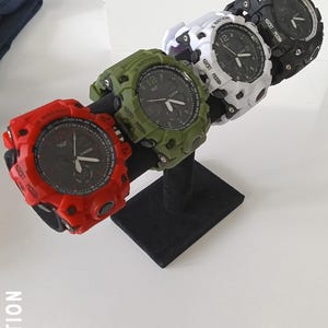 May include: Four digital watches in red, green, white, and black, displayed on a black stand. Each watch has a round face with a black dial and white numerals. The watches have a rugged, sporty design.