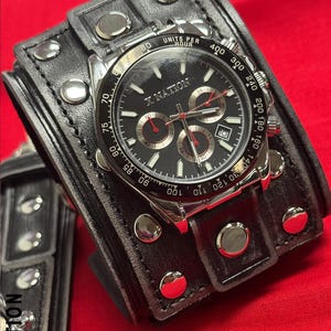 May include: A black wristwatch with a silver bezel and a black face, featuring red accents and the text "X NATION." The watch is set on a wide, black leather cuff with silver studs. The watch displays multiple subdials.
