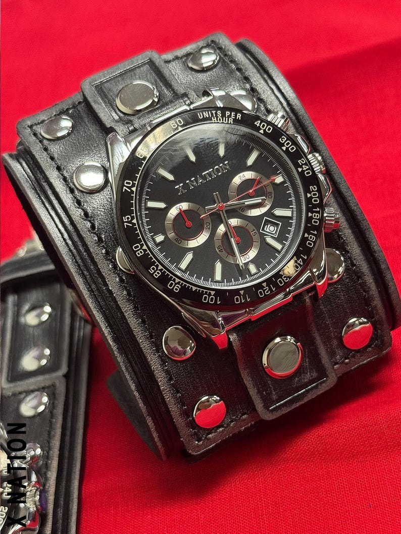 May include: A black wristwatch with a silver bezel and a black face, featuring red accents and the text "X NATION." The watch is set on a wide, black leather cuff with silver studs. The watch displays multiple subdials.