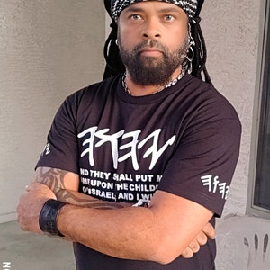 May include: A person wearing a black t-shirt with white Hebrew text and the words "AND THEY SHALL PUT M...ISRAEL, AND I W". The person is also wearing a black and white headscarf and a black wristband.