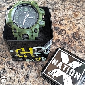 May include: An olive green digital watch with a black face, displayed in a black tin. The watch has multiple buttons and a textured band. The tin has the text "X NATION" and a stylized logo.