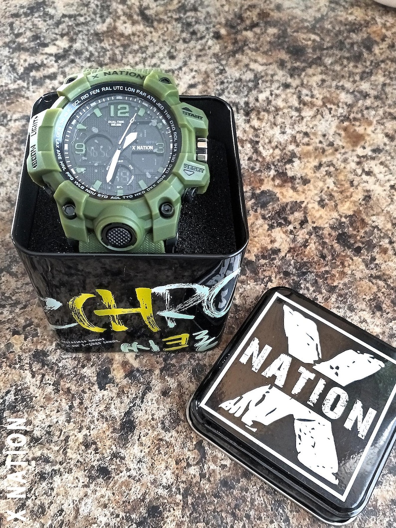 May include: An olive green digital watch with a black face, displayed in a black tin. The watch has multiple buttons and a textured band. The tin has the text "X NATION" and a stylized logo.