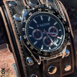 May include: A black-faced wristwatch with the text "X NATION" is attached to a wide, dark brown leather cuff. The watch has silver accents and red sub-dials. The cuff is secured with silver rivets and a buckle.
