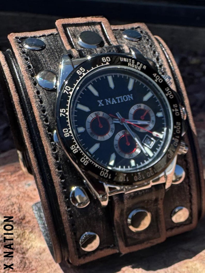 May include: A black-faced wristwatch with the text "X NATION" is attached to a wide, dark brown leather cuff. The watch has silver accents and red sub-dials. The cuff is secured with silver rivets and a buckle.
