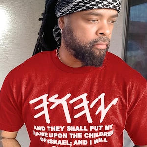 May include: Red t-shirt with white Hebrew text and the English text "AND THEY SHALL PUT MY NAME UPON THE CHILDREN OF ISRAEL; AND I WILL BLESS THEM. NUMBERS 6:27." The person is wearing a black and white patterned head covering.