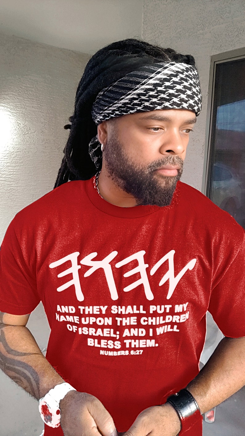 May include: Red t-shirt with white Hebrew text and the English text "AND THEY SHALL PUT MY NAME UPON THE CHILDREN OF ISRAEL; AND I WILL BLESS THEM. NUMBERS 6:27." The person is wearing a black and white patterned head covering.