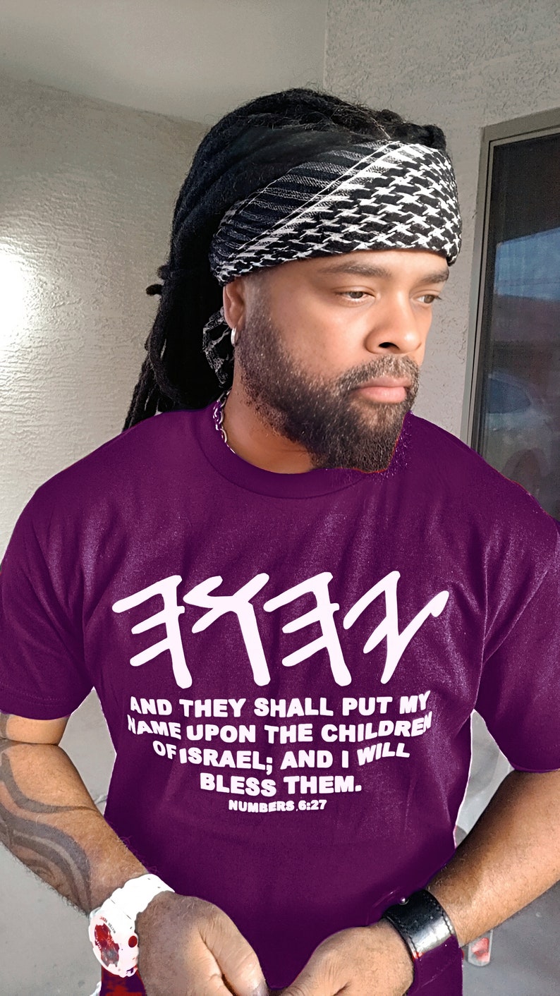 May include: A purple t-shirt with white Hebrew text and the quote "AND THEY SHALL PUT MY NAME UPON THE CHILDREN OF ISRAEL; AND I WILL BLESS THEM. NUMBERS 6:27." The person is wearing a black and white patterned head covering.