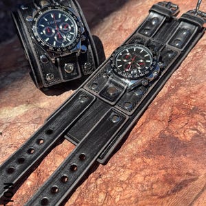 May include: Two black leather wrist cuffs with silver-toned watch faces. The watches have black dials with red accents and silver-toned bezels. The cuffs are riveted and have adjustable straps. The text "X NATION" is visible.