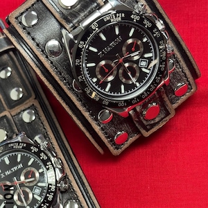 May include: Two wristwatches with black faces and silver-tone cases are displayed. The watches have multiple sub-dials and are set on wide, distressed leather bands with silver studs. The watch face reads "X NATION". The background is a solid red.