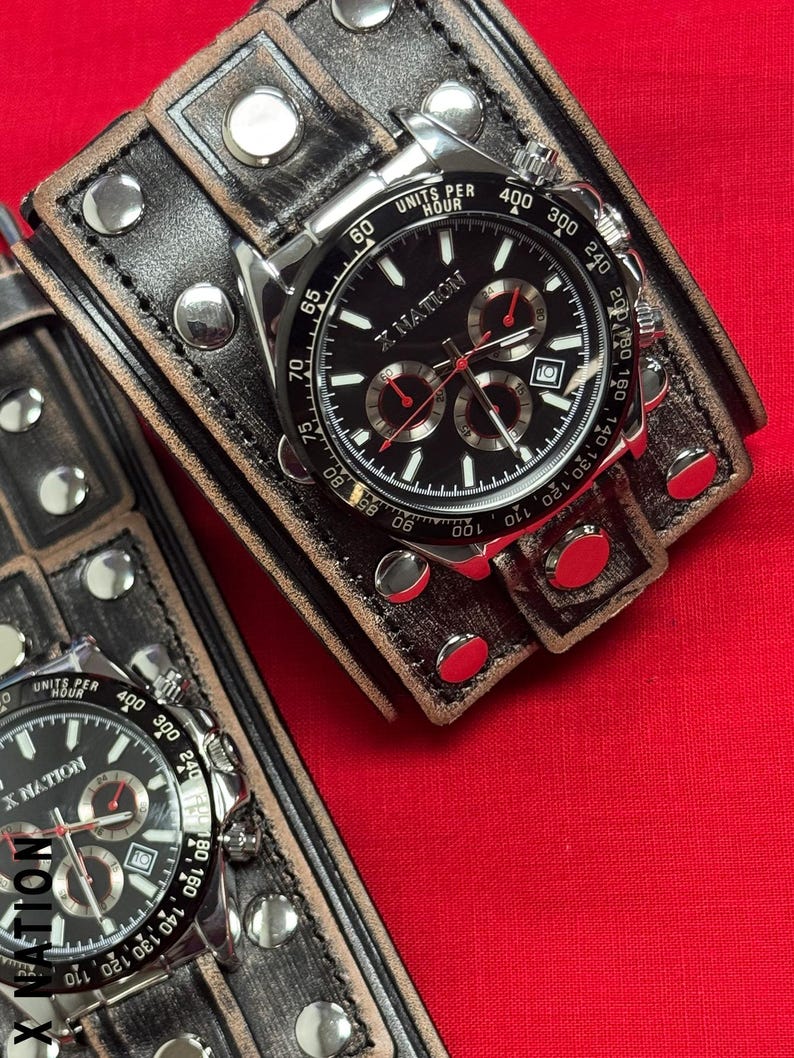 May include: Two wristwatches with black faces and silver-tone cases are displayed. The watches have multiple sub-dials and are set on wide, distressed leather bands with silver studs. The watch face reads "X NATION". The background is a solid red.