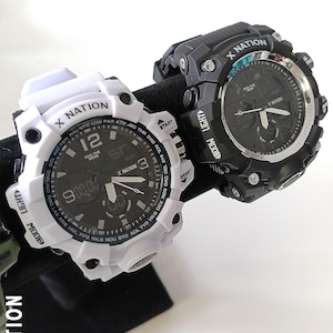 May include: Two digital watches with the text "X NATION" on the face and bezel. One watch is white with a black face, and the other is black with a black face. Both watches have multiple buttons and digital displays.